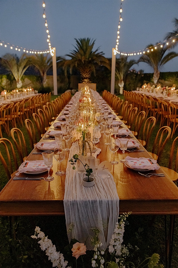 Reception tablescape with a long banquet table wedding setup, white runner, taper candles and florals under string lights on a twilight lawn with palm trees