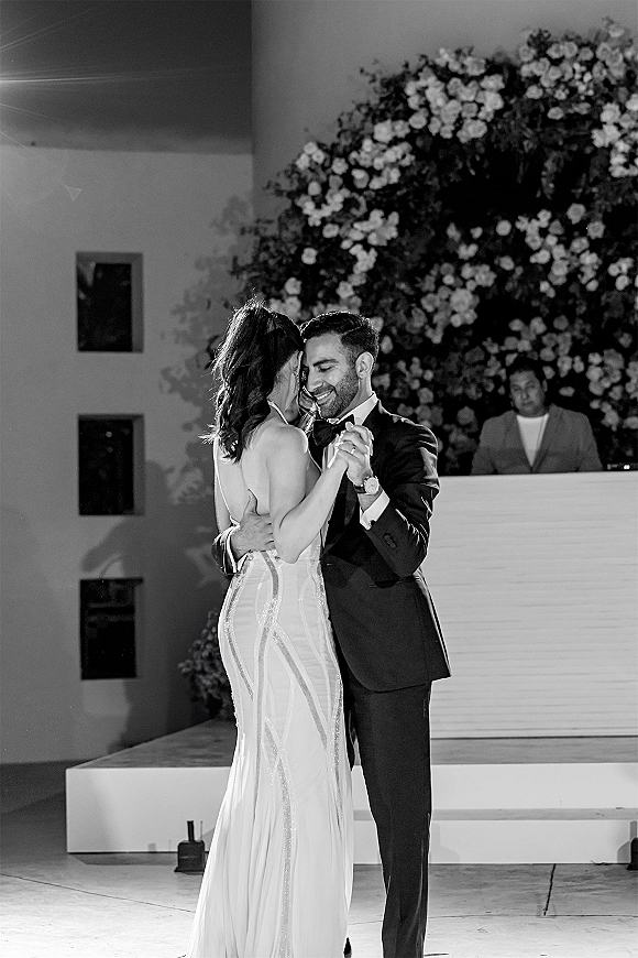 First dance as bride in beaded wedding gown and groom in tuxedo hold close on a stage, with a floral wall backdrop and DJ booth behind