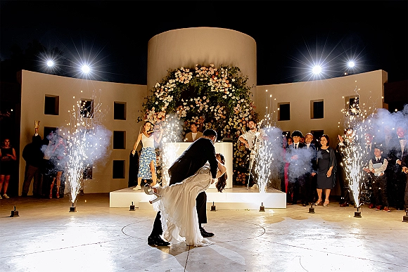 First dance moment as bride and groom dip for a kiss amid cold spark fountains and smoke on a stage under string lights at night