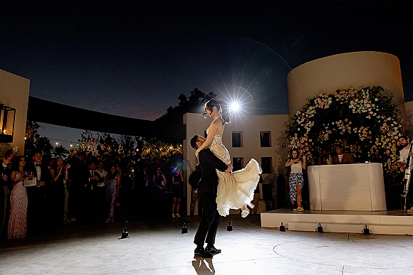 First dance moment as groom lifts bride in a strapless wedding dress under string lights, spotlighted in an outdoor courtyard with guests cheering