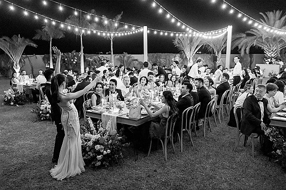 Wedding reception toast as a bridesmaid speaks under string lights, guests in tuxedos seated at candlelit banquet tables on a palm-lined lawn at night