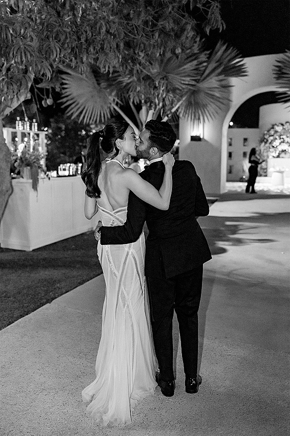 Wedding kiss as bride in a low-back gown and groom in a black suit embrace on a palm-lined outdoor walkway under night lights