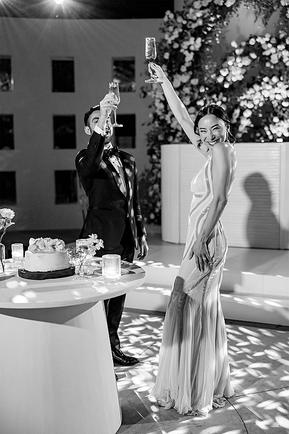 Wedding toast as bride in beaded gown and groom in tux raise champagne flutes by a small cake on stand with candlelit florals and floral wall backdrop