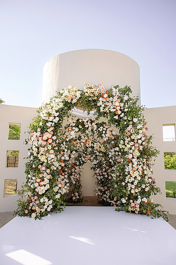 Wedding ceremony arch with roses and greenery, draped fabric accent, set in an outdoor courtyard against a white stucco wall and blue sky