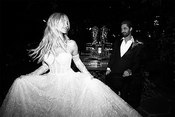 Couple portrait in black and white of bride in off-the-shoulder lace gown and groom in tux by champagne tower under garden string lights at night