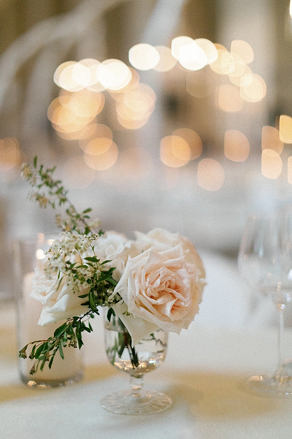 Wedding centerpiece with blush roses and greenery in a glass goblet vase, glowing by candlelight with bokeh string lights behind