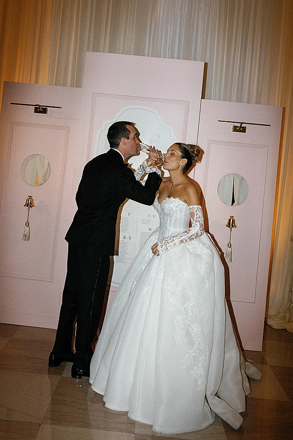 Champagne toast moment as bride and groom sip from flutes, her off-the-shoulder lace gown against pink backdrop panels on the dance floor