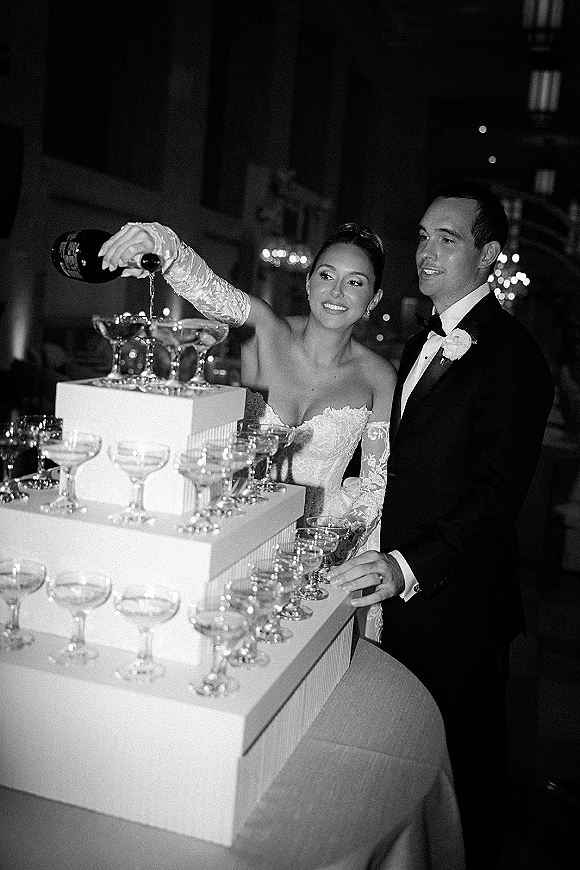 Champagne tower pour as bride in lace gloves and groom in tuxedo fill coupe glasses beneath chandeliers in a dim reception hall