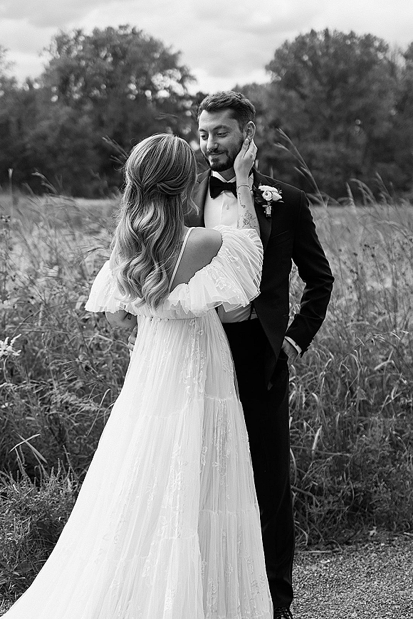 Couple portrait in a meadow, bride touching groom’s face in an off-the-shoulder lace gown as he wears a tuxedo with boutonniere
