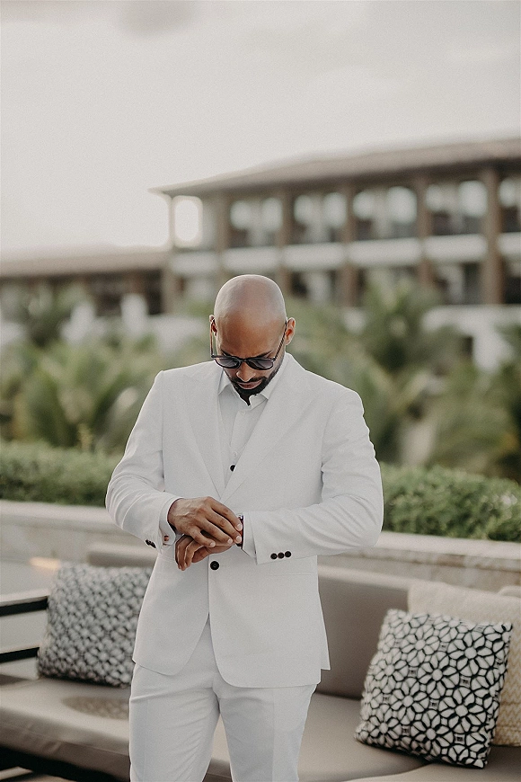 Groom portrait in a white wedding suit adjusting his wristwatch, wearing sunglasses on a modern patio with palm trees and lounge seating