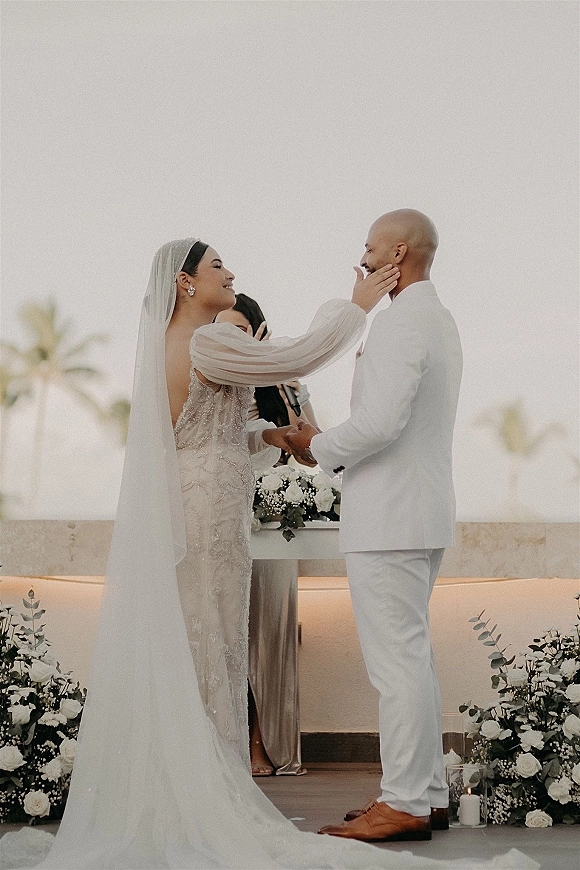 Wedding vows on an outdoor wedding vows terrace as bride touches groom’s face, beaded gown and veil, palm trees and candles behind
