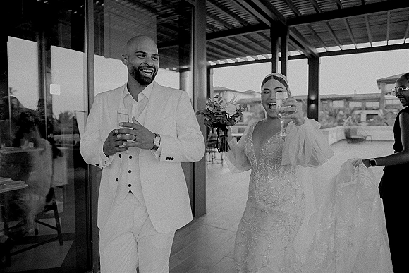 Reception entrance as bride and groom walk in laughing, she holding a champagne flute and bouquet on a covered patio with glass doors.
