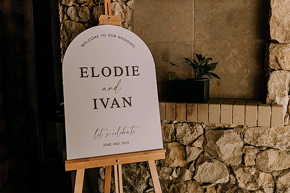 Wedding welcome sign with arched sign board on a wooden easel, calligraphy and serif lettering beside a potted plant by a stone fireplace wall