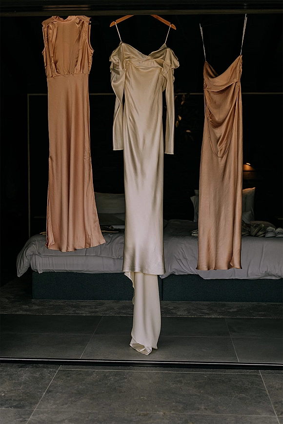 Wedding dress hanging above a bed, a satin wedding dress on a hanger beside champagne bridesmaid dresses against a dark wall