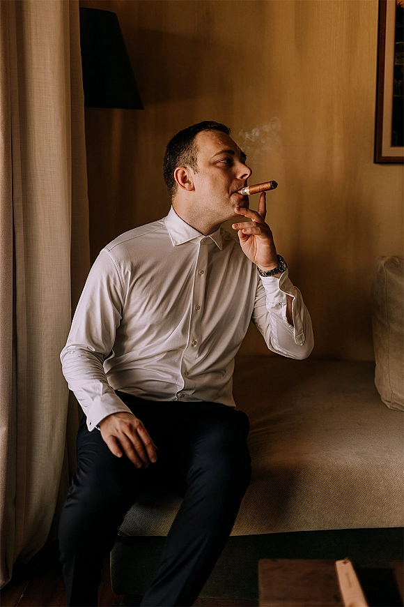 Groom portrait of a man smoking a cigar in a white dress shirt, seated on a sofa in a wood-paneled living room with window light