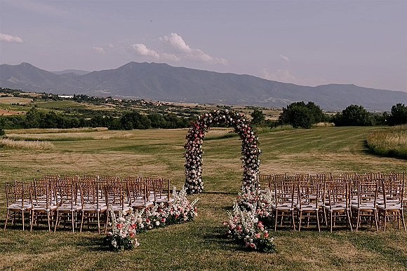 Outdoor ceremony setup with a floral ceremony arch and aisle flowers, chiavari chairs on a grassy field with mountain views under clouds