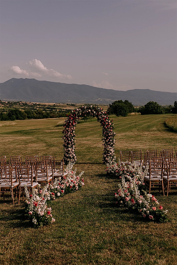 Ceremony setup with outdoor wedding ceremony floral arch and rose-lined aisle, wooden Chiavari chairs facing mountains under clouds