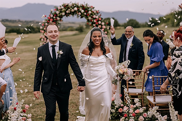 Recessional moment as bride and groom walking aisle under a circular floral arch, guests cheering amid rose petals on a mountain lawn