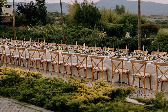 Reception tablescape with an outdoor wedding reception table set on a long banquet table, taper candles and floral centerpieces under string lights in a garden setting
