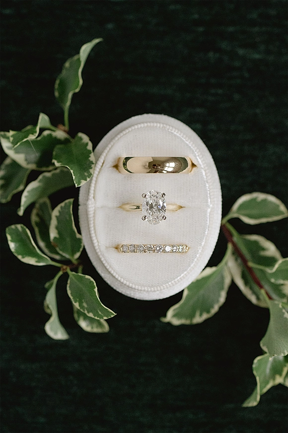 Wedding rings in a velvet ring box, featuring an oval diamond ring with diamond band on dark fabric with greenery accents