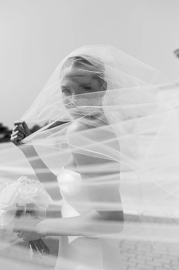 Bridal portrait of a bride with veil draped over her face, holding a rose bouquet in a strapless wedding dress outdoors under open sky