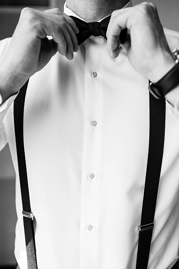 Groom getting ready, tying a black bow tie over a white dress shirt with suspenders and wristwatch in a softly blurred room