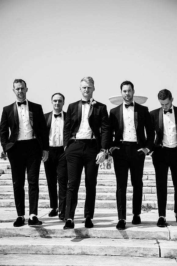 Groomsmen portrait of men in black tuxedos with bow ties and boutonnieres walking down stone steps near a fountain under open sky