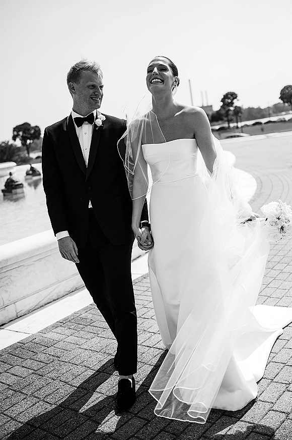 Wedding couple portrait of bride and groom walking hand in hand, laughing as her veil blows, beside a stone balustrade and water feature