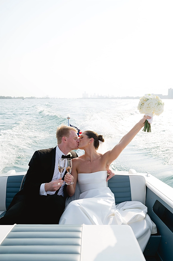Wedding kiss portrait of newlyweds on boat, bride holding bouquet and champagne flutes as ocean wake trails behind under open sky