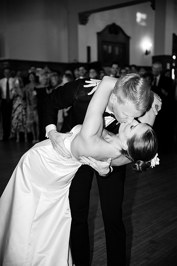 Wedding kiss portrait of groom dipping the bride in a dip kiss wedding photo on a wood dance floor, guests behind them in a reception hall