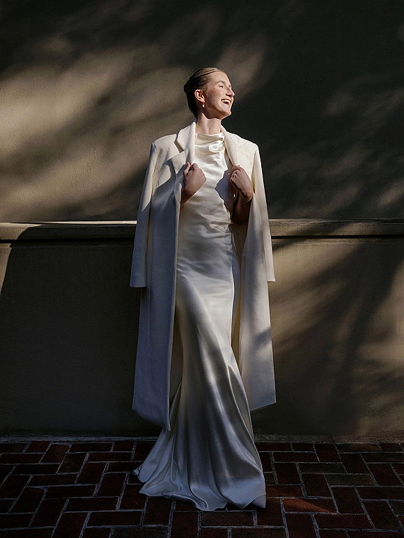 Bridal portrait of a bride in a modern bridal look wearing a satin wedding dress and long white coat by a stucco wall with shadows