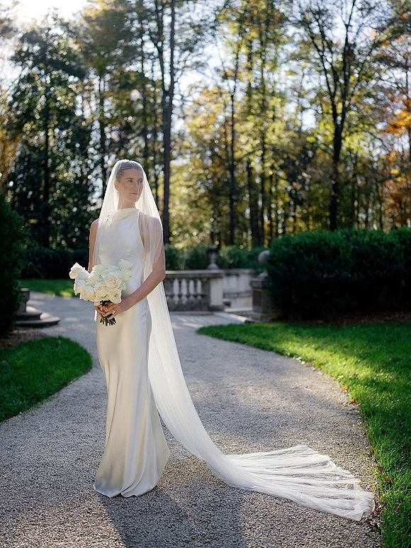 Bridal portrait of a bride holding bouquet of white roses, wearing a long veil and wedding dress on a sunlit garden path with trees