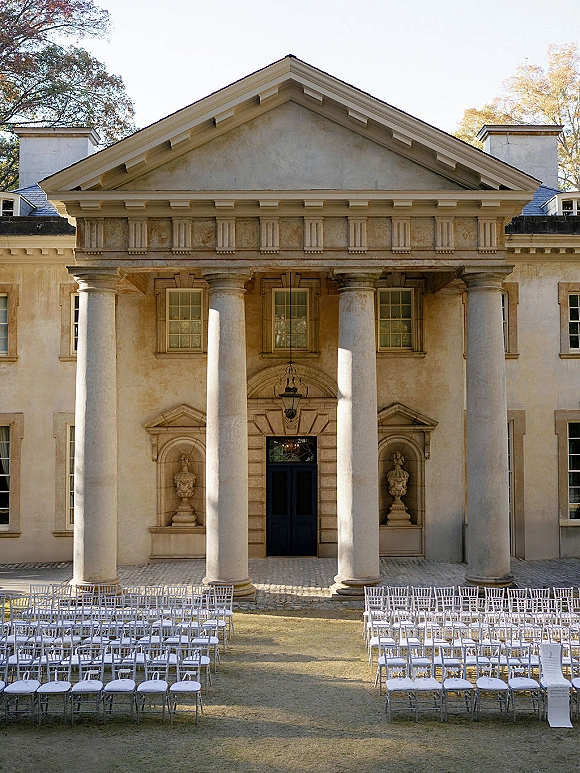 Ceremony setup with outdoor ceremony seating in neat rows of white chairs on a lawn before a neoclassical building with tall columns