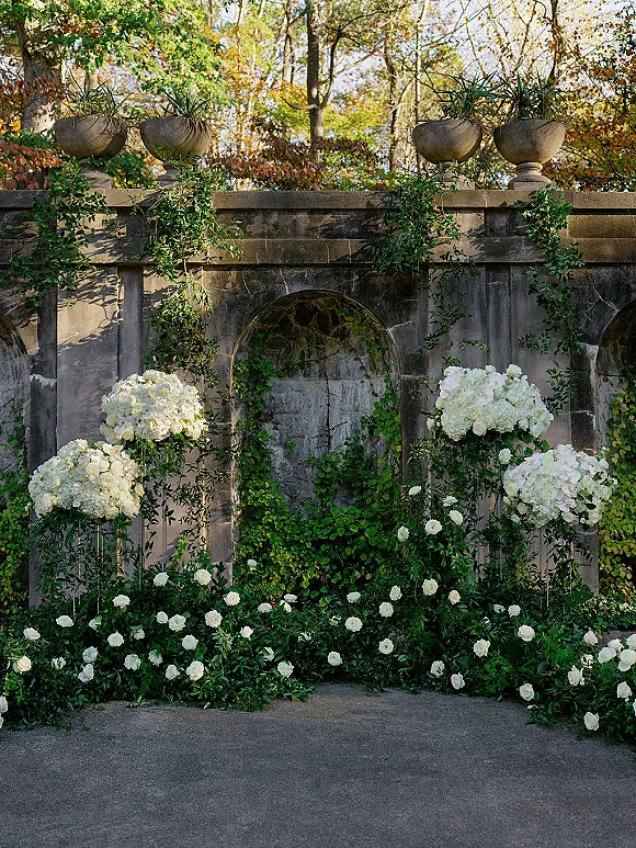Wedding ceremony backdrop with white rose arrangements and greenery garland against a stone wall in an outdoor garden courtyard setting