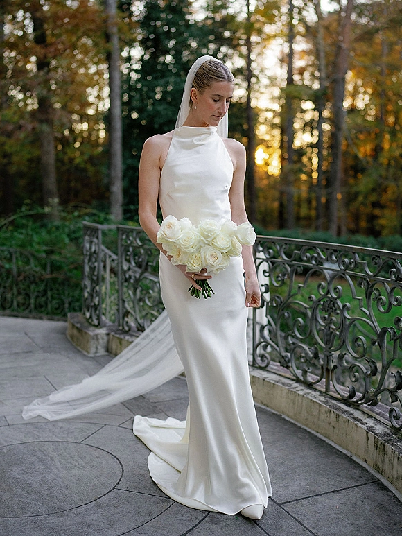Bridal portrait of a bride in a satin wedding dress holding a white rose bouquet on a terrace with wrought iron railing at sunset light