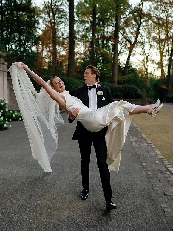 Couple portrait of groom carrying laughing bride, bridal veil blowing, on a tree-lined driveway beside a stone wall and lawn