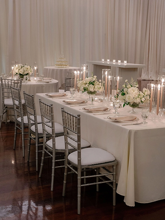 Reception tablescape with wedding head table decor featuring white linens, taper and votive candles, white florals, and chiavari chairs by draped walls