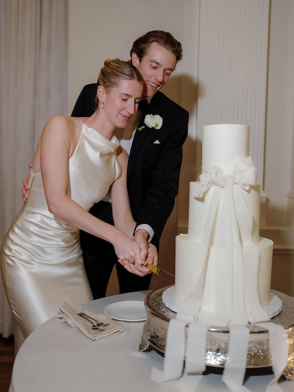 Cake cutting as bride in satin reception dress and groom in black tux slice a white tiered wedding cake on a silver stand in draped reception room