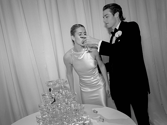 Champagne toast moment as groom in tuxedo feeds bride in satin slip dress a coupe beside a champagne tower and draped curtain backdrop