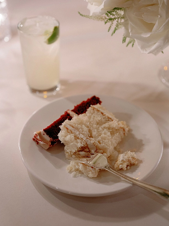 Wedding cake slice on a white plate with dessert fork, cake crumbs, and a drink glass with lime wedge on a floral-detailed tablecloth