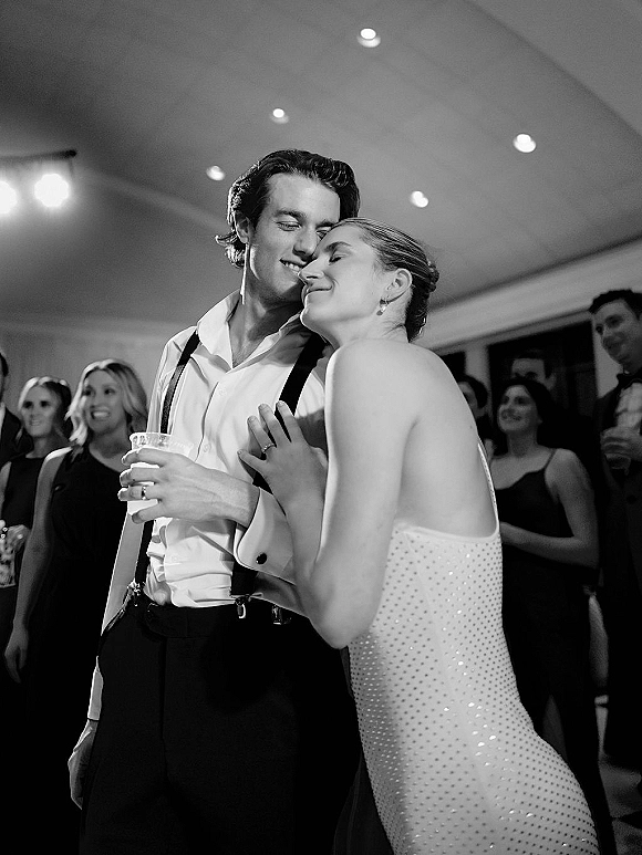 Wedding dance moment as bride in a strapless dress leans on groom in suspenders holding a cocktail, guests and ceiling lights behind