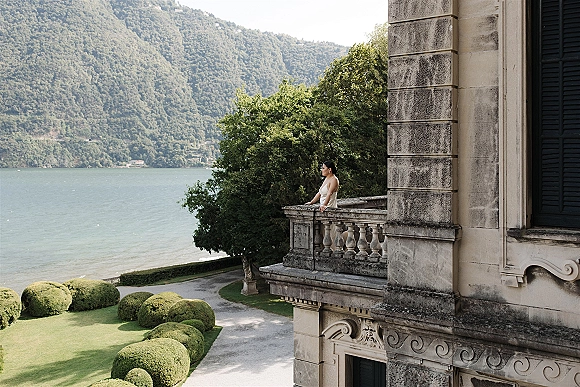 Bridal portrait of a bride on balcony in a long wedding dress, leaning on the balustrade with a lake and mountains beyond