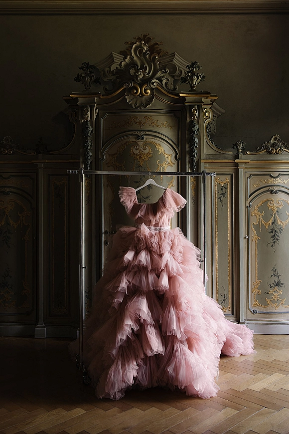 Wedding dress in blush pink hanging on a rack, with tulle ruffles and embellished belt against an ornate carved wardrobe in dim light