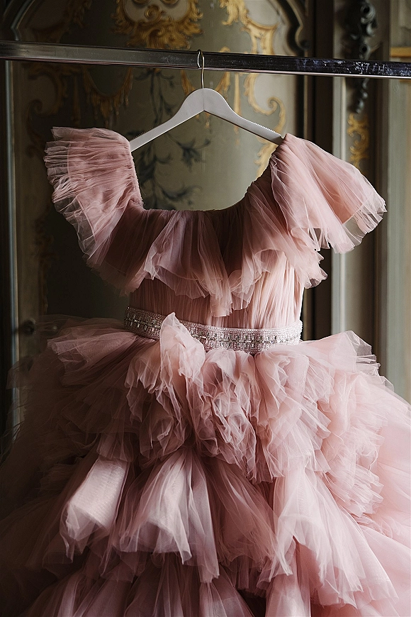 Wedding dress in soft blush with off-shoulder ruffles and beaded belt hanging on a rack in a vintage room with ornate wallpaper