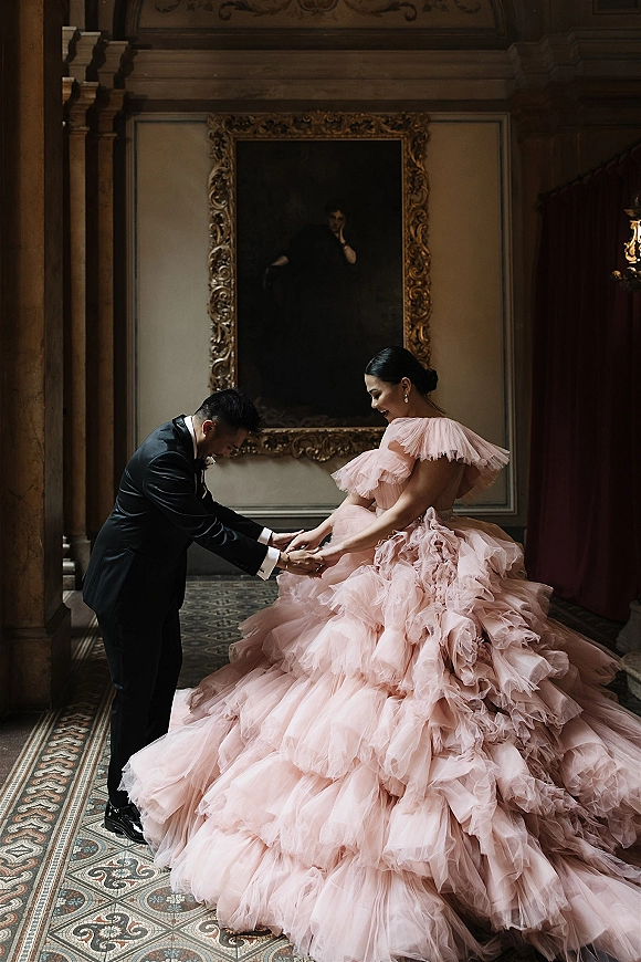 Couple portrait of groom kissing bride’s hand as they hold hands, her blush pink tulle ball gown flowing in an ornate interior with columns
