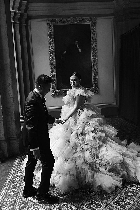 Couple portrait in a black and white wedding portrait style, bride in tulle ruffled ball gown holding groom’s hand in a grand hallway