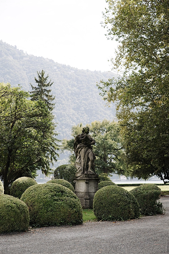 Garden statue stone garden sculpture on a pedestal beside manicured shrubs, with a gravel path leading to trees, lake, and mountains