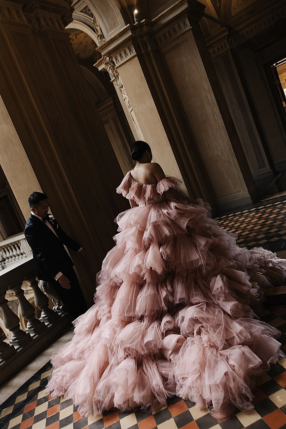 Couple portrait of bride in blush tulle ball gown with long train and groom in black tuxedo walking through a grand stone hallway