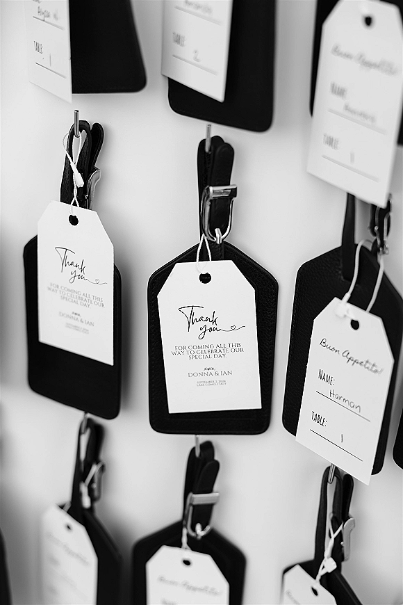 Wedding escort cards styled as luggage tag escort cards with calligraphy text and metal hooks on black backers against a white wall