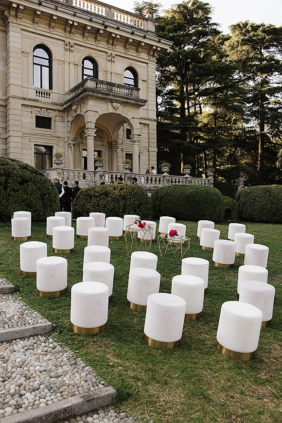 Ceremony seating with white velvet ottomans on gold bases in a circular layout, with pink florals on side tables on a manicured lawn by a stone villa